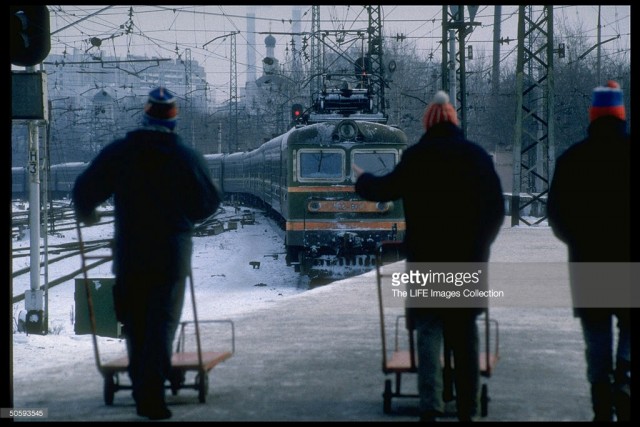 Trio of empty luggage-cart pushing men poised on platform facing arriving train at the Kursky railway station, Moscow (February 19, 1992).jpg